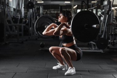 Atletik kadın modern spor salonunda halter çömeliyor. Güç motivasyonunu ve sağlıklı yaşam tarzını gösteriyor.