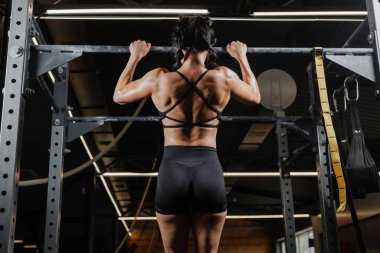 Spor salonunda barfiks çeken atletik bir kadın güçlü sırt kaslarını güçlendiriyor spor yapma gücünü ve motivasyonunu ön plana çıkarıyor.