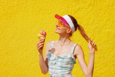 Güneş gözlüklü ve güneşli neşeli bir kadın sıcak yaz gününde canlı sarı duvara gülümsüyor.