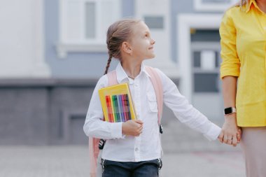 Okul çantalı ve kırtasiyeli mutlu öğrenci kız annemle el ele tutuşuyor, okula döndüğü ilk gün için heyecanlı..