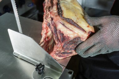Kasap, kurutulmuş çiğ eti metal testereyle dilimlerken paslanmaz çelik masaya güvenlik eldiveni takıyor.