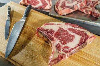 Kurumuş pirzola bifteğinin üst görüntüsü. Temiz çalışma alanındaki mutfak bıçaklarıyla kesme tahtasına yerleştirilmiş mermer yağlı biftek.