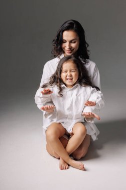 Anne ve kızı birbirine uyumlu kıyafetlerle oturmuş gülümsüyor ve stüdyo ortamında el ele oynuyorlar.