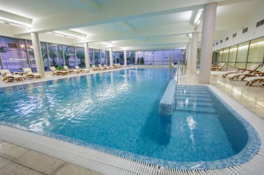 Parlak, modern bir otel kaplıcasında ya da sağlık merkezinde mavi suyu ve şezlongları olan geniş bir yüzme havuzu.