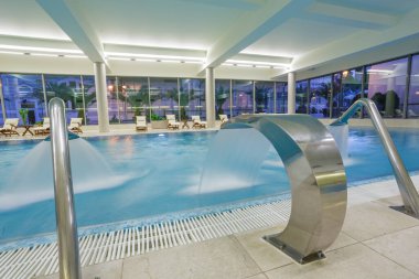 Şelale özellikli geniş bir spa havuzu ve modern otel sağlık merkezinde dinlendirici şezlonglar.