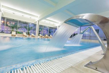 Kapalı bir spa havuzu, su taşması ve dinlendirici şezlonglar, modern, aydınlık bir dinlenme tesisi.