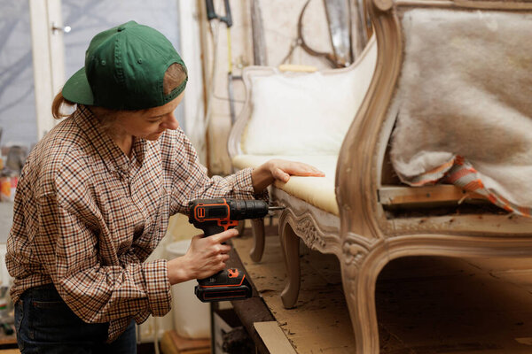 Женщина ремесленник реставрации винтажного кресла с беспроводной дрель в мастерской, подробный процесс ремонта мебели