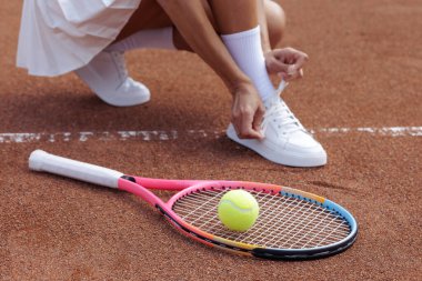 Bir kadın oyuncunun kil yüzeyde raket ve topun yanına spor ayakkabısı bağlaması, antrenman, spor kıyafeti ve spor kıyafeti içeriği için ideal..