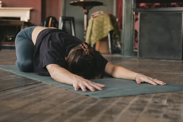 Full-body photo of a female yogi resting in child\'s pose (Balasana) with her arms extended during her vinyasa flow yoga practice alone and wearing sportswear indoors
