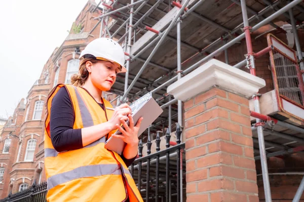 Chartered civil engineer lady writing on a tablet with electronic pen, hard hat and orange high visibility vest, inspection survey, touchscreen, technology and innovation in construction site