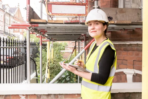 Smiley civil engineer woman inspecting a site with a tablet and electronic pen, wears hard hat and yellow high visibility vest, innovation and technology in construction, outdoors, looking at camera