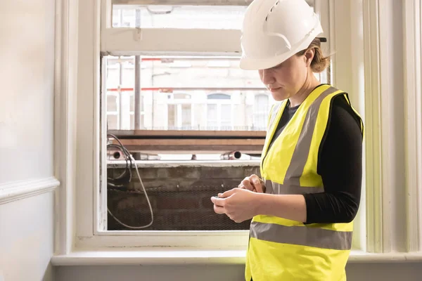 Civil engineer woman using a calculator in front of a window in a construction site, wears yellow high visibility vest, ppe, personal protective equipment, indoor site supervision