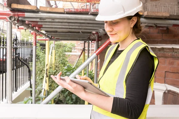 Smiley female civil engineer taking notes on a tablet with an electronic pen, wears hard hat and yellow personal protective equipment, innovation and technology in construction, outdoors, side view