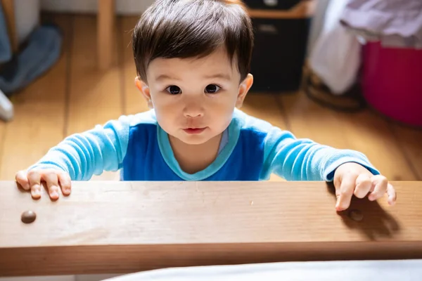A multiracial Malaysian and Spanish baby wearing blue pajamas staring into the camera with mucus under his nose because he is sick and unwell. Touching wooden furniture with his hands.