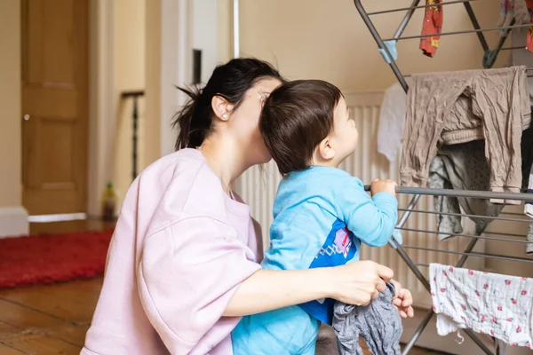 An unrecognizable new mother is trying to hang out the laundry and wet clothes while her son, a male infant, gets in the way indoors. Real mixed race family at home in pajamas.