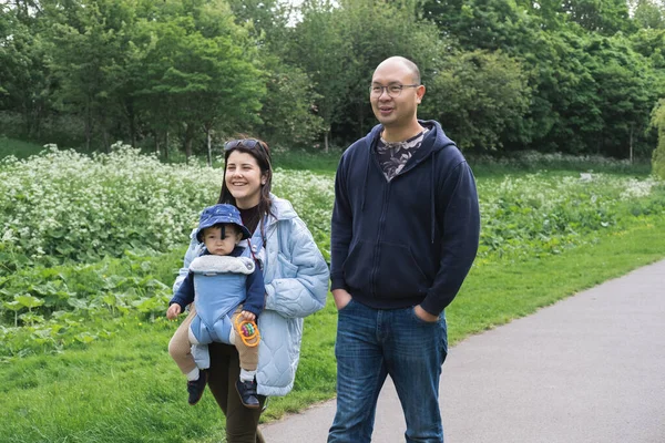 A multicultural family walking on a footpath in Figgate Park in Edinburgh in spring. The mother is carrying her infant son with a baby carrier. The baby wears a misplaced hat and is uncomfortable.