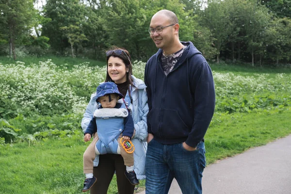 A smiley multicultural family walking on a footpath in Figgate Park in spring season. The mother is carrying her infant son with a baby carrier. The baby wears a misplaced hat and carries a toy.