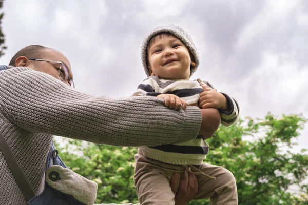 Yeni baba tatlı bebekle oynuyor, kaldırıyor, sevimli bebeği havaya fırlatıyor, sevgiyle gülen çocuğu kollarına alıyor, babalığın tadını çıkarıyor, ebeveynliğin tadını çıkarıyor. Çok ırklı, melez bir aile. Düşük açı görünümü.