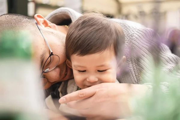 Dreamy picture of father with son, male infant sitting on his dad\'s legs with glasses in beer garden at pub in Edinburgh. Intimate caring moment. Bokeh with plant. Multicultural family, mixed race.
