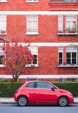 İngiltere 'nin Londra sokaklarında kırmızı ağaçlı kırmızı tuğlalı bir binanın önüne park edilmiş kırmızı küçük bir arabanın dikey fotoğrafı..