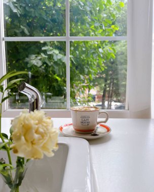 Pencerenin arkasında beyaz bir çiçek olan bir fincan kahve..