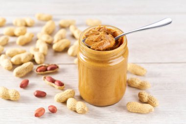 peanut paste in an open jar and peanuts in the peel scattered on wooden table.
