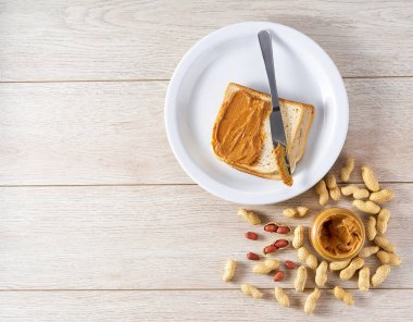 peanut butter in an open jar on a white wooden background, next to peanuts scattered.