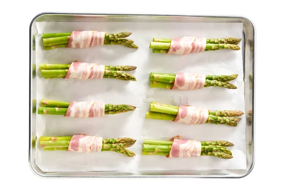准备烘焙开胃菜时 用熏肉包裹的绿色芦笋捆在烤盘上 与白色背景隔开 — 图库照片
