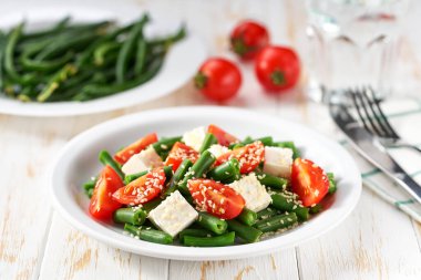 Beyaz ahşap bir masada domates, feta ve susam tohumlu bir kase salata. Beyaz tabakta yeşil fasulye ve domates salatası..