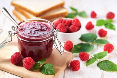 Beyaz ahşap bir masada cam bir kavanoz ahududu reçeli. Ev yapımı ahududu reçeli, taze böğürtlen ve mutfak masasında yeşil yaprak..