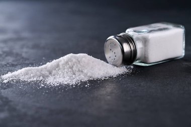 Siyah bir mutfak masasına tuzlanmış tuzluk ve dökülen deniz yığını seçici bir odak noktası. Tuz karıştırıcı ve ters dönmüş tuz..