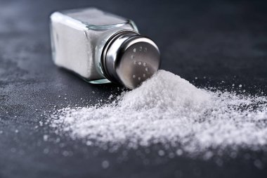 Tuzluk ve dökülmüş tuz kristalleri siyah bir masanın üzerine, seçici bir odak noktası. Tuzluktan bir yığın tuz, aşırı tuz alımı kavramı..