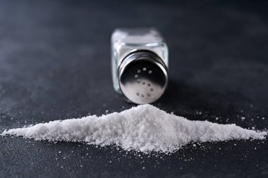 Tuzluk ve dökülmüş tuz kristalleri siyah bir masanın üzerine, seçici bir odak noktası. Tuzluktan bir yığın tuz, aşırı tuz alımı kavramı..