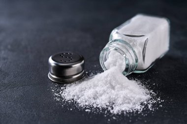 Siyah bir mutfak masasına tuzlanmış tuzluk ve dökülen deniz yığını seçici bir odak noktası. Tuz karıştırıcı ve ters dönmüş tuz..