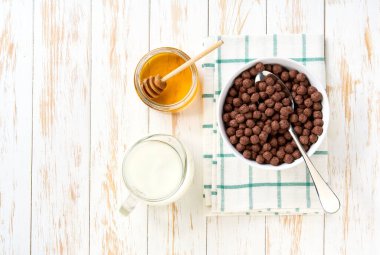 Beyaz ahşap bir masa üzerinde sütlü ve ballı seramik bir kasede çikolatalı mısır topları. Çikolatalı mısır köfteli sağlıklı kahvaltı.