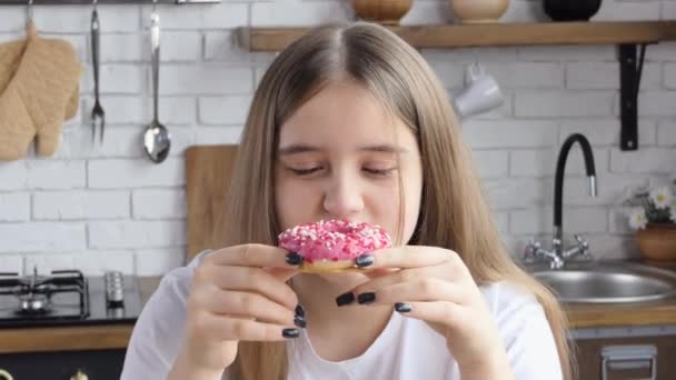 クローズアップのポートレート 幸せな美しい若い女の子が食欲をそそるピンクのドーナツを噛み 彼女の唇を舐め 笑います 台所の女の子は陽気にお菓子を食べます — ストック動画