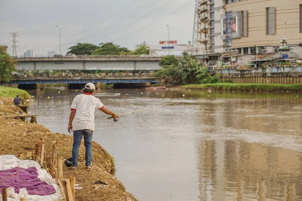 Jakarta, Endonezya - 11 Aralık 2022: Jakarta 'nın batı kanalı üzerinde öğleden sonra balık tutan şapkalı, beyaz tişörtlü ve kot pantolonlu bir adamın arka görüntüsü.