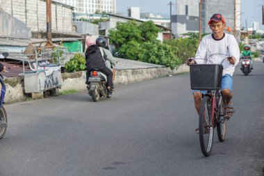 Jakarta, Endonezya - 1 Nisan 2023: Güneşli bir öğleden sonra asfalt bir yolda bisiklet süren uzun kollu gömlek, şapka ve şort giyen yaşlı bir adamın portresi.