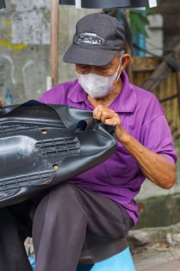 Jakarta, Endonezya - 28 Mart 2024: Motosiklet koltuğu tamircisi olarak çalışan şapka ve maske takan yaşlı bir adam yeni bir motosiklet koltuğu örtüsü takıyor.