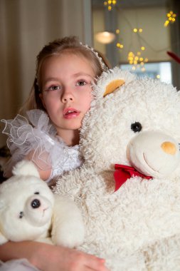 Kollarında oyuncak ayısıyla yerde oturan küçük bir kız..