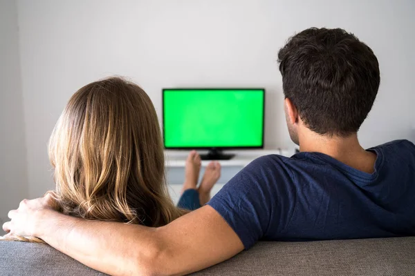 Back view of young family couple watching TV with chroma key while resting at home sitting on sofa in room.