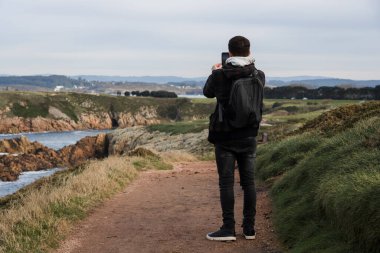 Bir uçurumun kenarında bulutlu bir gökyüzüne karşı dururken cep telefonuyla manzarayı fotoğraflayan genç bir adamın arka görüntüsü.