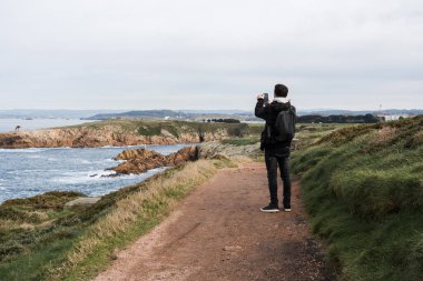 Bir uçurumun kenarında bulutlu bir gökyüzüne karşı dururken cep telefonuyla manzarayı fotoğraflayan genç bir adamın arka görüntüsü.