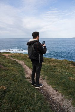 Bir uçurumun kenarında bulutlu bir gökyüzüne karşı dururken cep telefonuyla manzarayı fotoğraflayan genç bir adamın arka görüntüsü.