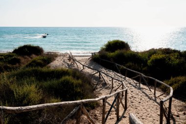 Tahta tahta kaldırım sahile çıkıyor. Odun, korkuluk, kum çimi. Arka planda okyanus ve mavi gökyüzü. Agrigento, Sicilya, İtalya