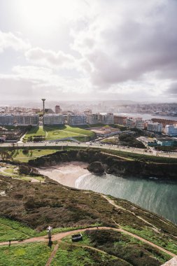 A Coruna, İspanya 'nın yüksek açılı hava manzarası, kıyı şeridini, kumlu sahilini, yerleşim binalarını ve uzaktaki ikonik Torre de Hercules' i gösteriyor. Çevrede yeşil parklar ve dolambaçlı yollar var. Gökyüzü bulutlu.