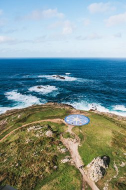 A Coruna, İspanya 'nın yüksek açılı hava manzarası, kıyı şeridini, kumlu sahilini, yerleşim binalarını ve uzaktaki ikonik Torre de Hercules' i gösteriyor. Çevrede yeşil parklar ve dolambaçlı yollar var. Gökyüzü bulutlu.