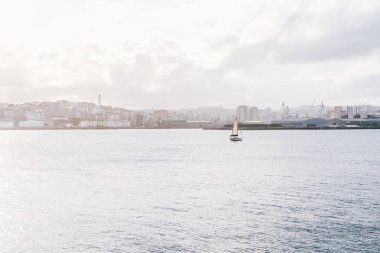 A Coruna, İspanya 'nın sakin sularında süzülen sakin bir yelkenli sahnesi. Görüntü, sahil şeridi boyunca yumuşak ışıklandırma ve endüstriyel yapılarla huzurlu bir deniz atmosferi yakalıyor.