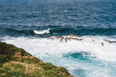 A Coruna, İspanya 'nın kıyı kayalıklarından Atlantik Okyanusu manzarası, kayalık kıyı şeridi boyunca canlı yeşil çimenler ve bitki örtüsü içerir. Açık mavi gökyüzü ve dağınık bulutlar sahnenin doğal güzelliğini arttırıyor, tema için mükemmel.