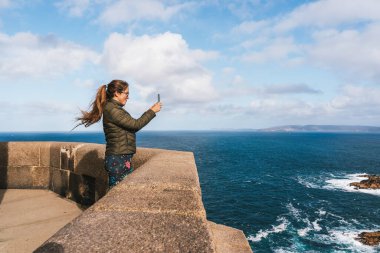 A young woman standing at a coastal viewpoint, taking a selfie with a smartphone. She is wearing a green jacket and patterned leggings, with long hair blowing in the wind.  clipart